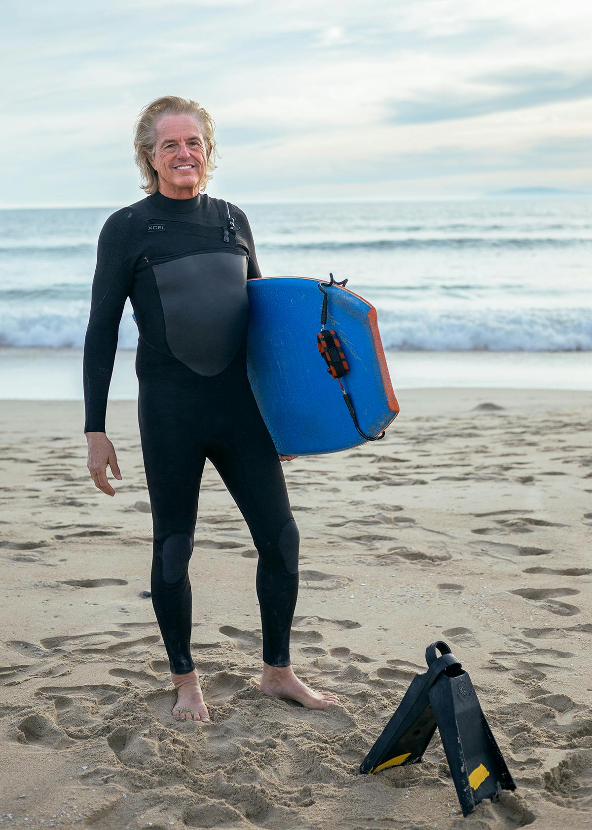 Man standing in wetsuit with boogie board and fins.